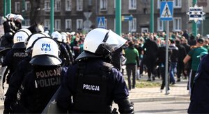 Kolorowe zdjęcie policjantów Oddziałów Prewencji Policji ubranych w kaski i wyposażonych w tarcze konwojujący kibiców.