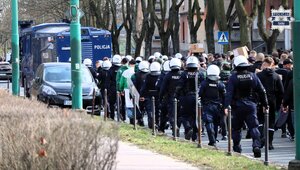 Kolorowe zdjęcie policjantów Oddziałów Prewencji Policji ubranych w kaski i wyposażonych w tarcze konwojujący kibiców.
