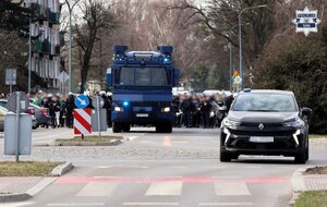 Kolorowe zdjęcie policjantów Oddziałów Prewencji Policji ubranych w kaski i wyposażonych w tarcze konwojujący kibiców. Na czele widoczny jest pojazd specjalny policji "armata wodna" oraz nieoznakowany radiowóz Policji.