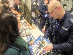 Umundurowany policjant wręczający uczestnikom gazetki promocyjne Policji.