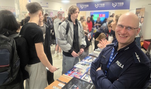 Uśmiechnięty policjant. W tle uczestnicy targów pracy.