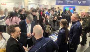 Umundurowani policjanci stojący przy stoisku promocyjnym Policji. W pobliżu duża grupa osób uczestniczących w targach pracy.