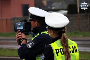 Umundurowany policjant drogówki wykonujący pomiar prędkości za pomocą laserowego miernika prędkości. Za nim stoi umundurowana policjantka drogówki.