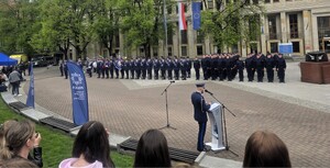 Umundurowany policjant stojący przy mównicy, w tle widoczni umundurowani policjanci stojący na placu.