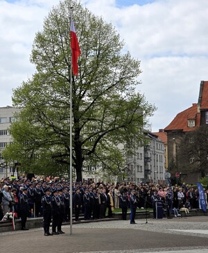 Umundurowani policjanci stojący w pobliżu masztu na której zawieszona jest flaga Rzeczpospolitej Polski.