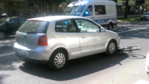 Samochód marki Volkswagen Polo stojący na jezdni. Z prawej strony pod pojazdem widoczny jest fragment roweru, natomiast w tle widoczny jest oznakowany radiowóz Policji.