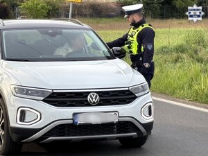 Umundurowany policjant Wydziału Ruchu Drogowego przeprowadza badanie kierującego samochodem marki Volkswagen.