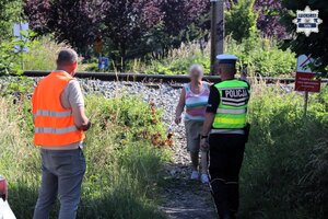 Pracownik Polskich Linii Kolejowych oraz umundurowany Policjant Wydziału Ruchu Drogowego podchodzą do kobiety, która przeszła przez torowisko w miejscu zabronionym.