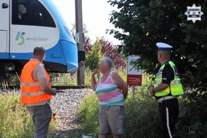 Pracownik Polskich Linii Kolejowych oraz umundurowany Policjant Wydziału Ruchu Drogowego rozmawiają z kobietą. W tle widoczne jest torowisko oraz przejeżdżający nim pociąg.