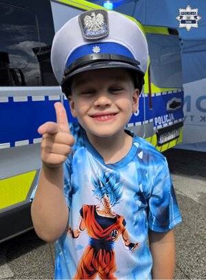 Chłopiec ubrany w policyjną czapkę stojąc na tle radiowozu Policji pokazuje gest kciukiem ręki.