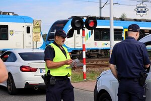 Umundurowany policjant oraz pracownik Straży Ochrony Kolei rozdają ulotki kierowcom. W tle widoczne jest torowisko, którym nadjeżdża pociąg.