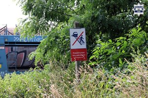 Znak stojący w pobliżu torów kolejowych zabraniający przejścia przez tory. W tle widoczne składy kolejowe.