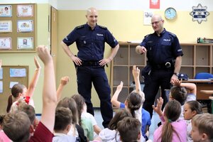 Dwóch umundurowanych policjantów stojących przed grupą siedzących na dywanie dzieci, które zgłaszają się przez podniesienie rąk.