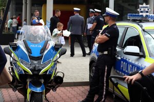 Umundurowani policjanci stojący obok oznakowanego motocykla oraz radiowozu policji.