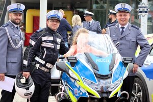 Dziewczynka siedząca na  oznakowanym motocyklu Policji. Obok stoją umundurowani policjanci.