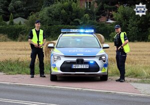 Umundurowani policjanci stoją obok oznakowanego radiowozu Policji.