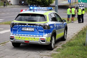 Oznakowany radiowóz Policji. W tle widoczny policjant oraz pracownicy zabezpieczenia przejazdu rowerowego ubrani w kamizelki odblaskowe.