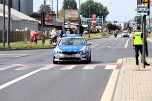 Oznakowany radiowóz Policji filtrujący drogę przed nadjeżdżającym peletonem kolarskim. W tle widoczny stojący policjant.