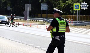 Umundurowany Policjant Wydziału Ruch Drogowego kontroluje ruch w rejonie oznakowanego przejścia dla pieszych. W tle widoczny przechodzący pieszy prowadzący rower na przejściu dla pieszych oraz stojący pojazd, które ustępuje pierwszeństwo przechodzącemu pieszemu.
