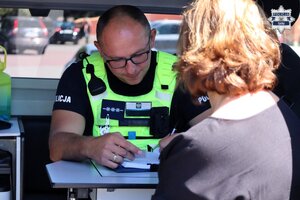 Umundurowany Policjant Wydziału Ruch Drogowego wypisuje mandat karny. W tle widoczna kierująca samochodem.