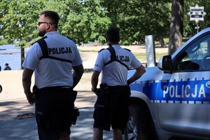 Umundurowani policjanci w umundurowaniu letnim stojący w pobliżu oznakowanego radiowozu. Spoglądają w dal w kierunku jeziora.