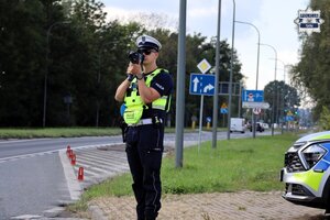 Umundurowany Policjant Wydziału Ruchu Drogowego stojący przy jezdni podczas pomiaru prędkości ręcznym miernikiem prędkości. W tle widoczny radiowóz Policji.