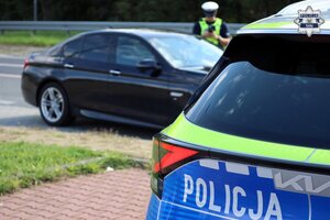 W tle oznakowanego radiowozu Policji widoczny policjant przeprowadzający kontrolę drogową.