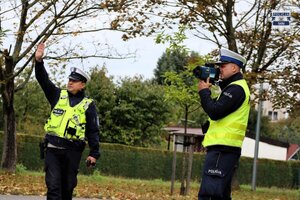 Umundurowani Policjanci Wydziału Ruchu Drogowego stojący przy jezdni, jeden wydaje polecenie do zatrzymania pojazdu przez podniesienie ręki do góry, drugi przeprowadza pomiar prędkości przy użyciu laserowego miernika prędości.
